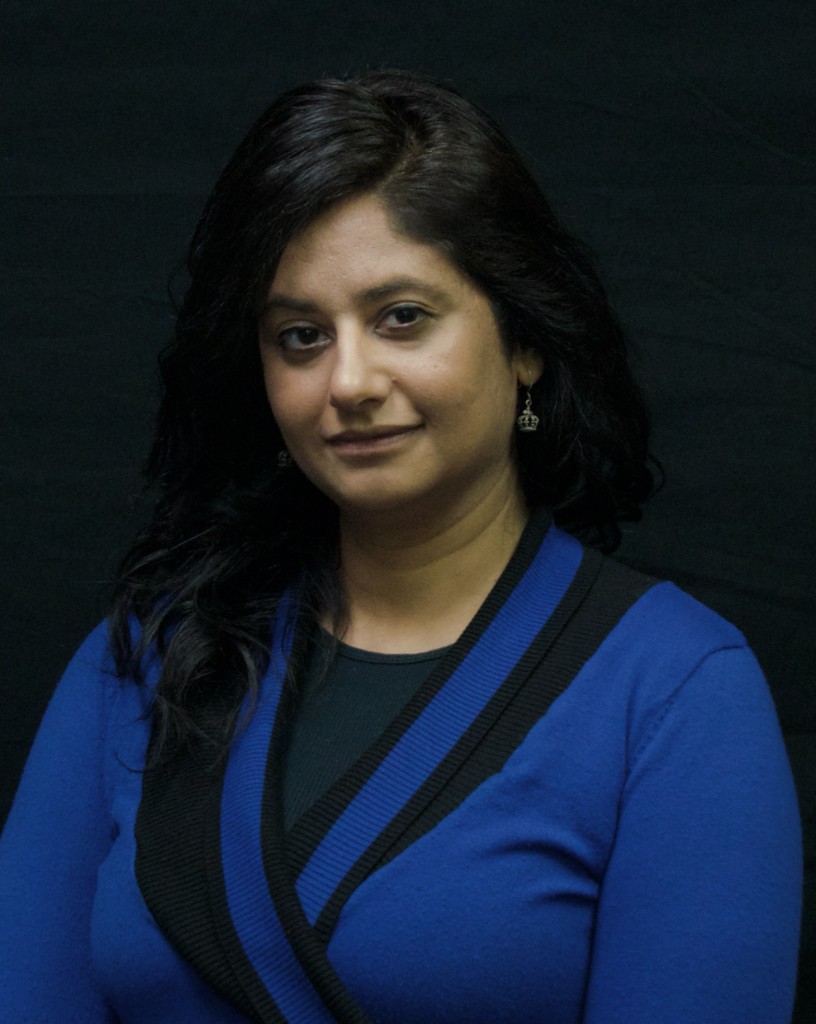 Professional headshot of Mandeep "Monica" Dhillon, Real Estate Agent, wearing a blue cardigan against a black background.